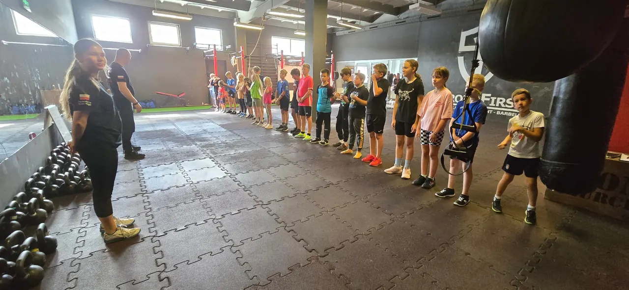 Dzieci ustawione w szeregu obok instruktorki, odważniki kettlebell i worek bokserski